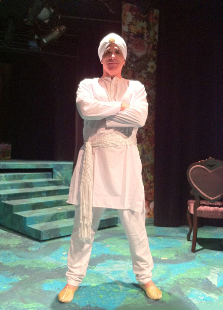 Person in white traditional attire with turban and sash on colorful stage with floral backdrop and chair.