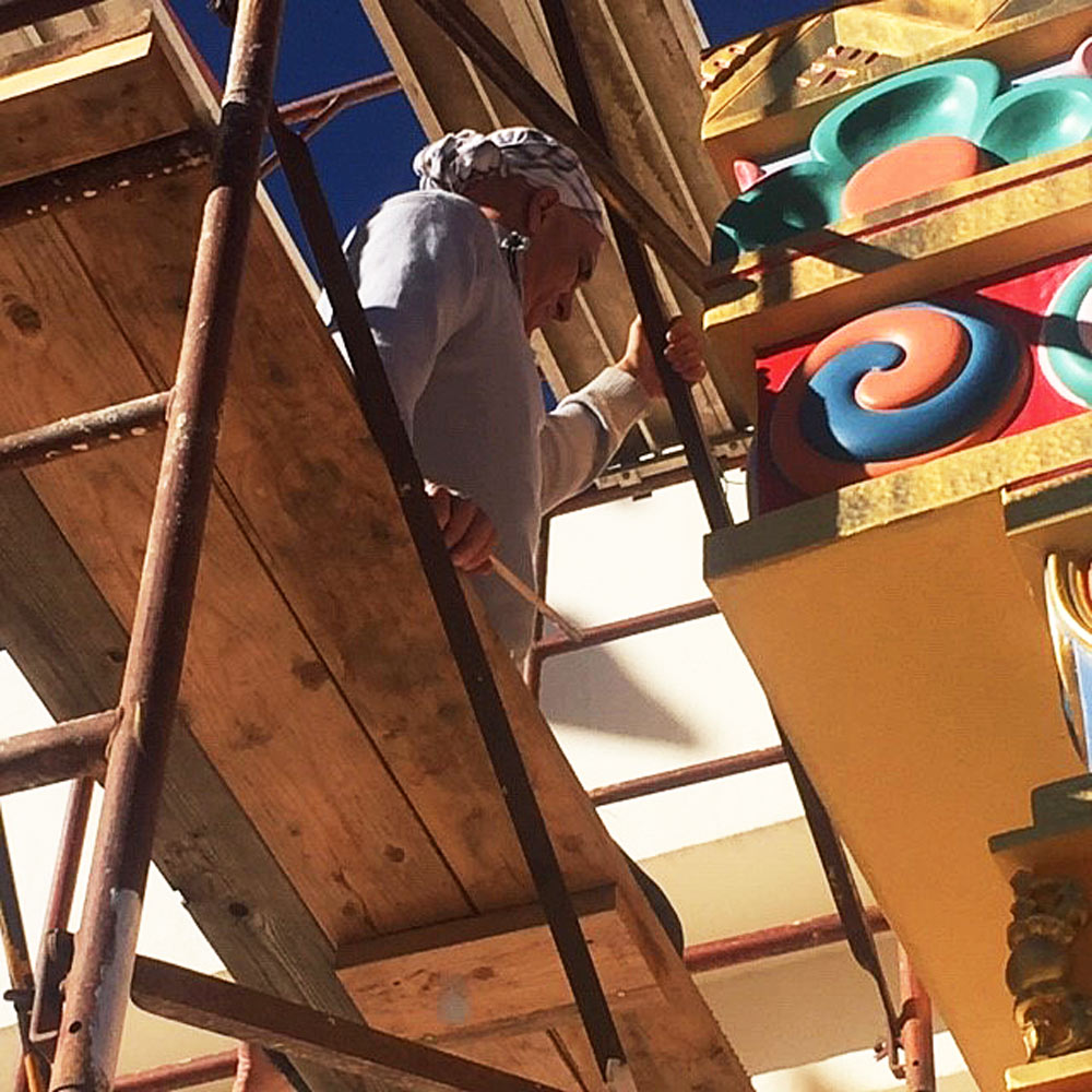 A ground-level view of artist, Ralph Williams (wearing a light blue sweater and a white & blue head-wrap) sitting on scaffolding and preparing to do touch-up painting work on the decorative elements (known as "tababs") on one of the four gates that accent The Great Stupa of Dharmakaya, a monument to human kindness.