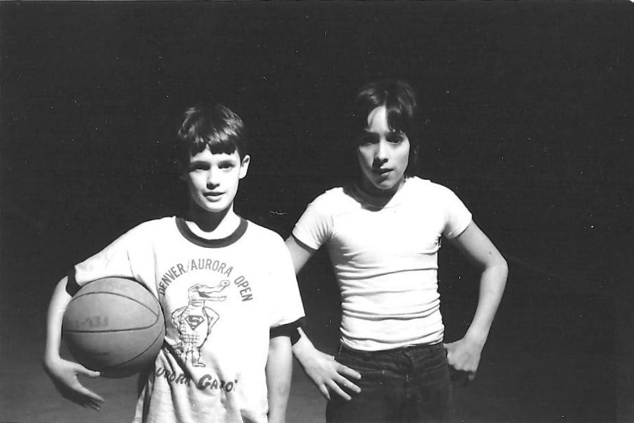 Two boys in dim light, one holding a basketball, both wearing T-shirts, standing side by side.