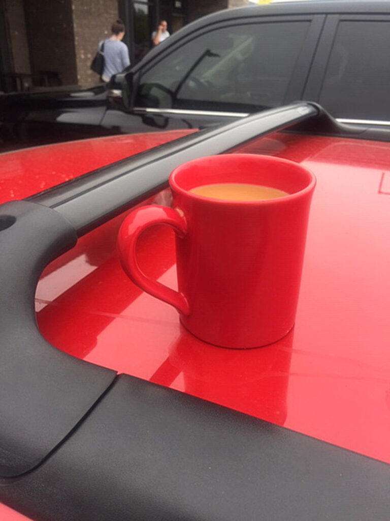 Red coffee cup with light brown beverage on red vehicle roof, black vehicle and building in background.