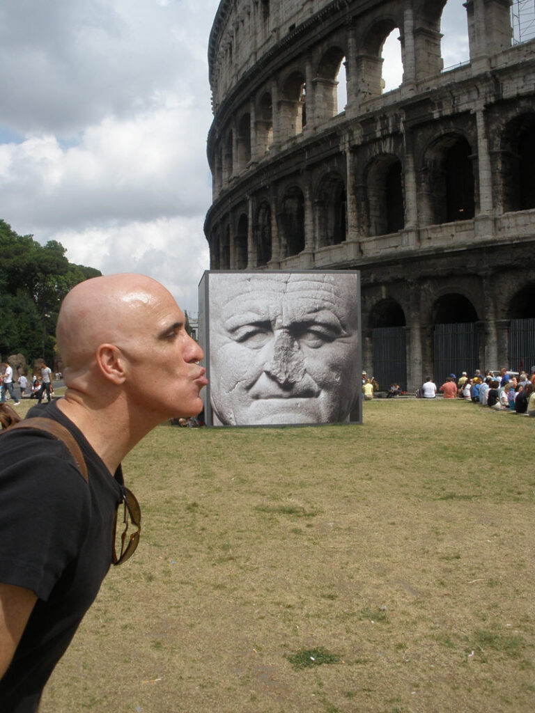 Person mimics kissing a large elderly man's face sculpture near the Colosseum in Rome, with tourists.