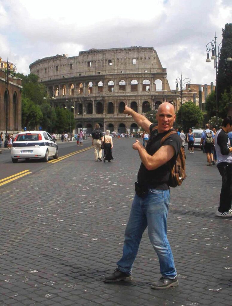 Man in casual attire with backpack points toward Colosseum in Rome on a cobblestone street.