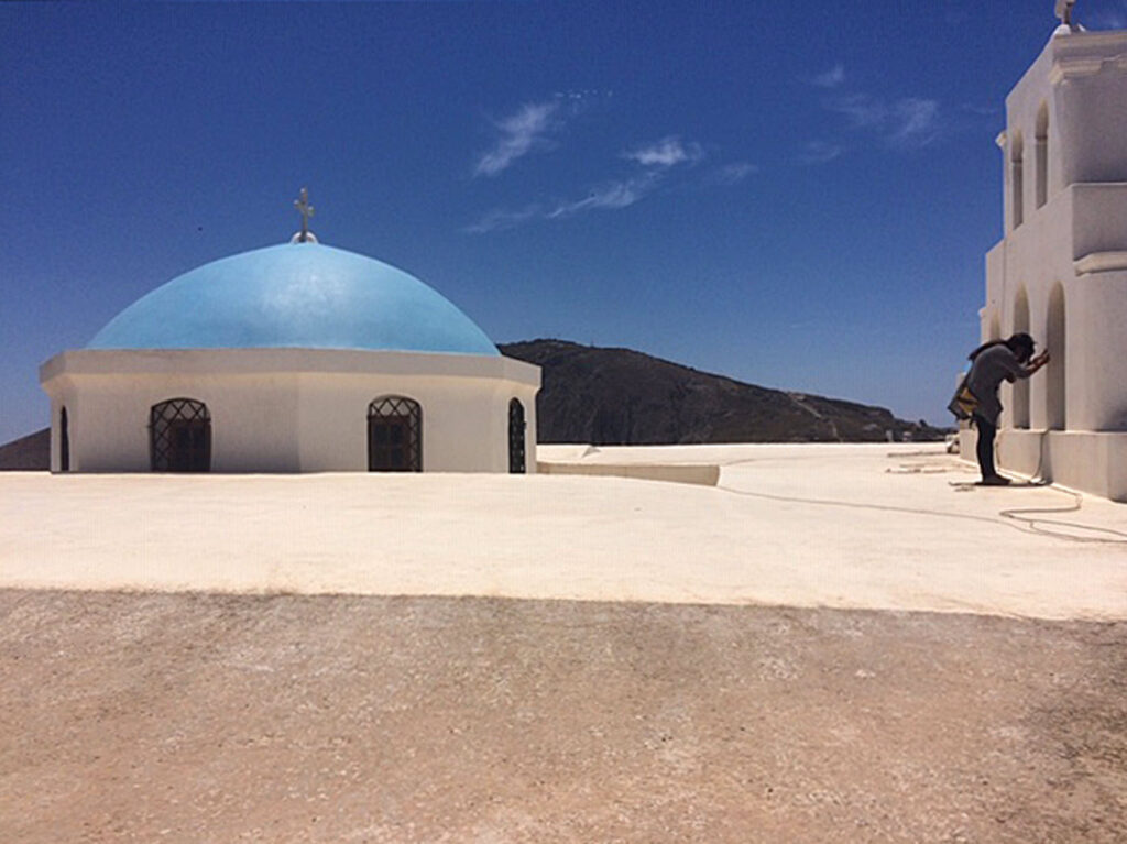 A classic blue dome with a small gold cross at the top accents a beige-concrete roof as a young woman (bent over at the waist) takes a photo through a window on a large gateway partition.