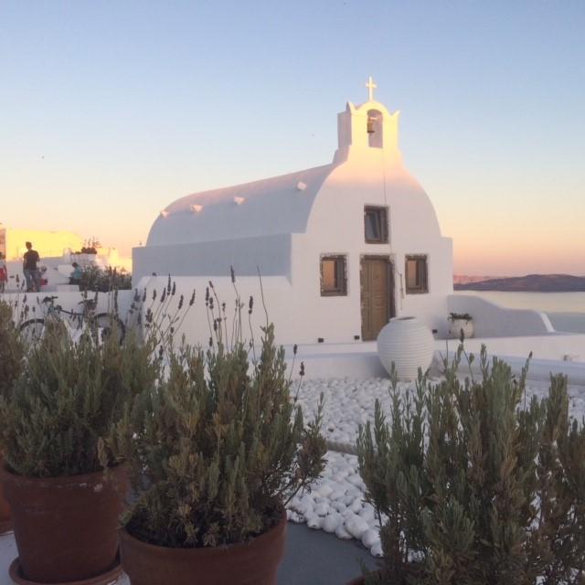 Whitewashed building with a domed roof and bell tower, potted plants, sea in background at sunset.