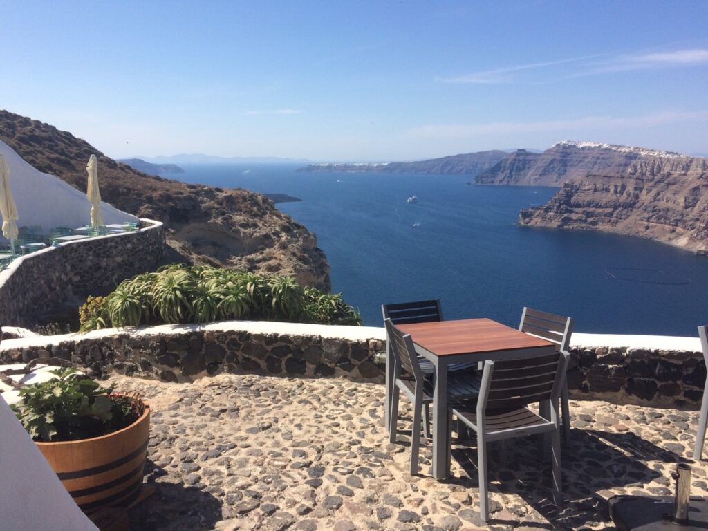 Stone-paved terrace with tables, chairs, greenery, white buildings, overlooking coastal cliffs and sea