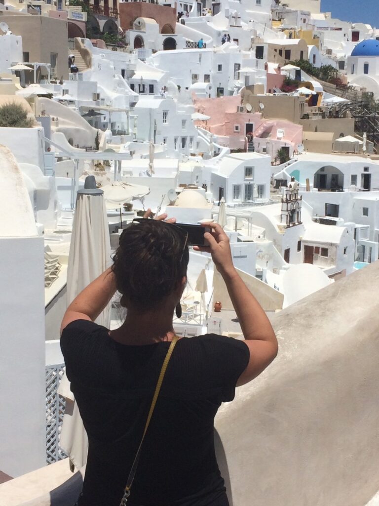 Person photographing Mediterranean-style hillside buildings with flat roofs and domes on a sunny day