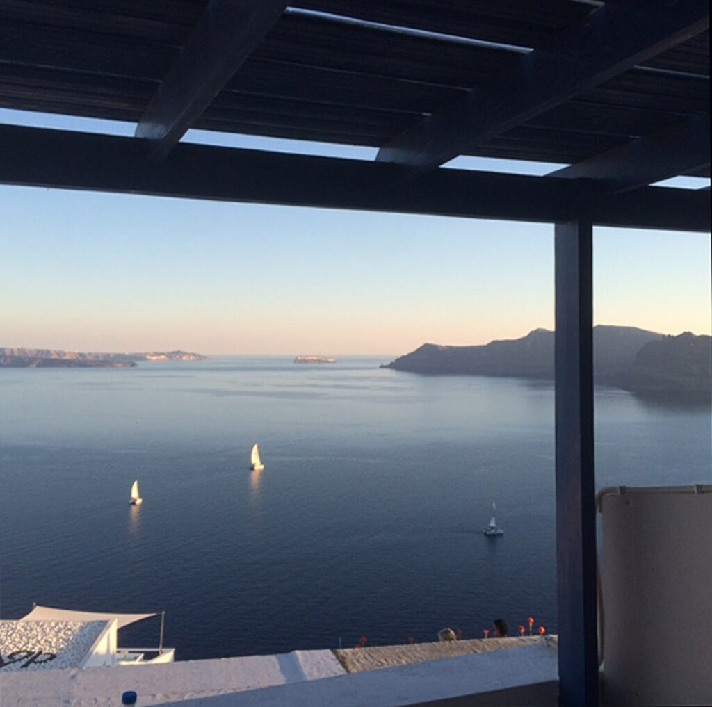 Seascape view with sailboats, distant coastline, islands, and the roof of a white building in foreground.