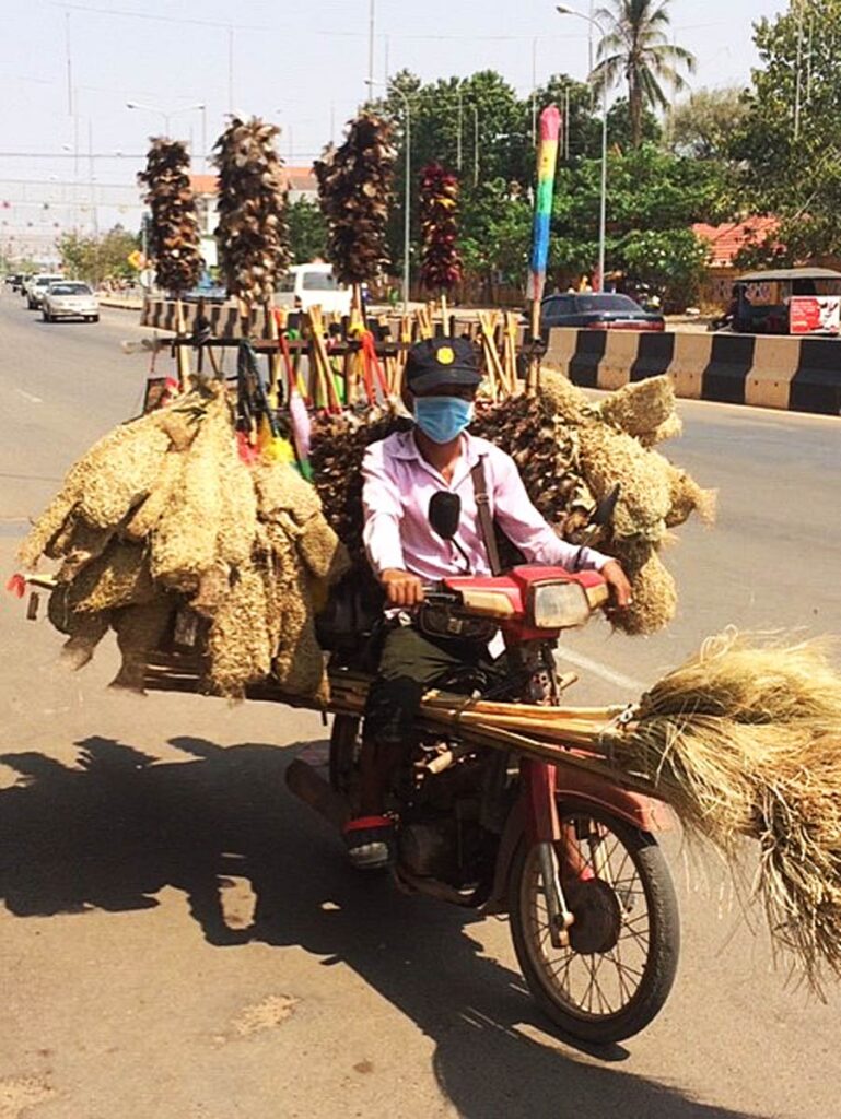 Person on motorbike carrying many brooms and cleaning tools, wearing protective gear on a city street.