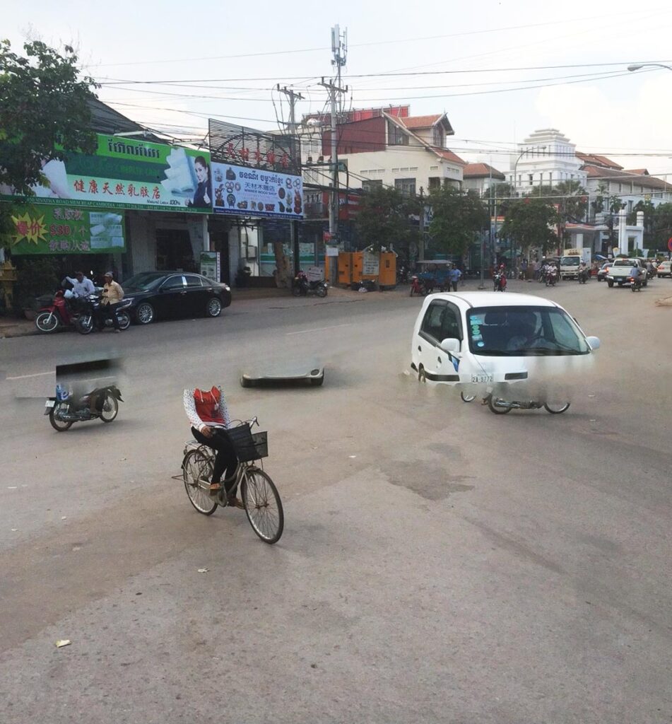 A camera glitch produces a surreal image of a wide intersection in Siem Reap, Cambodia with a headless person riding a bicycle and a whte van without wheels running into a motorscooter.