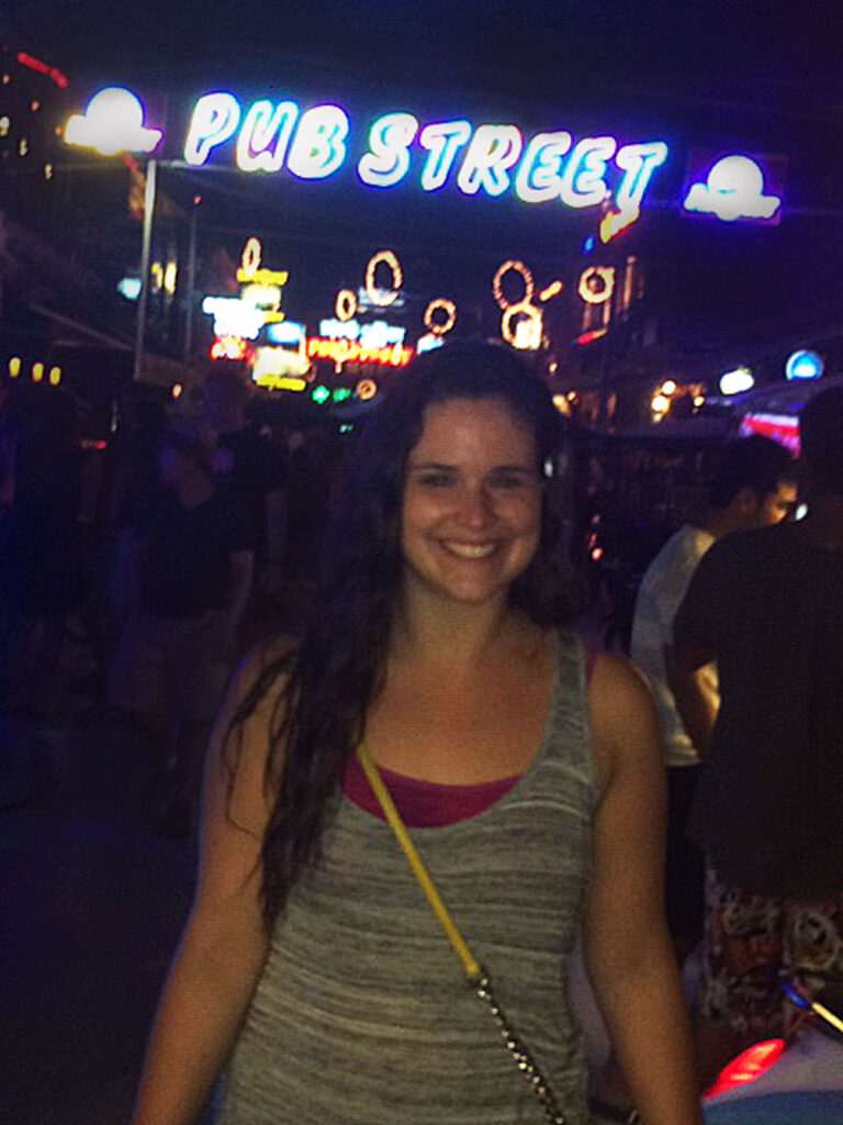 Woman standing on a busy, neon-lit Pub Street at night, wearing a sleeveless top and carrying a bag.