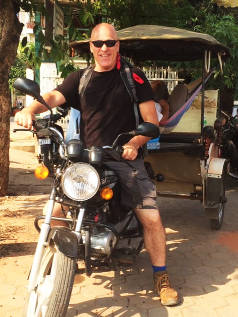 Person with sunglasses on motorcycle, parked on paved street, tuk-tuk and tree in the background.