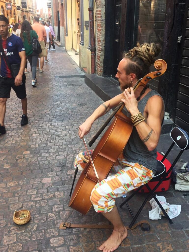 Street musician with dreadlocks and colorful shorts playing a cello barefoot on a cobblestone street