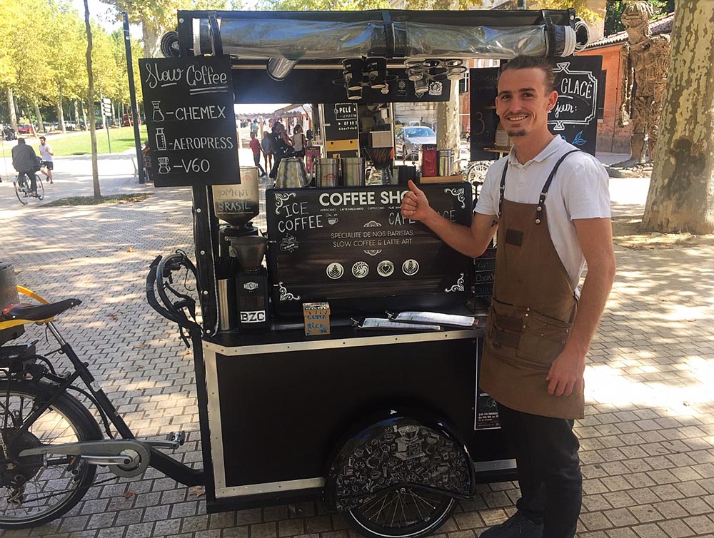 Person beside a mobile coffee cart offering various coffee options in an outdoor park setting.