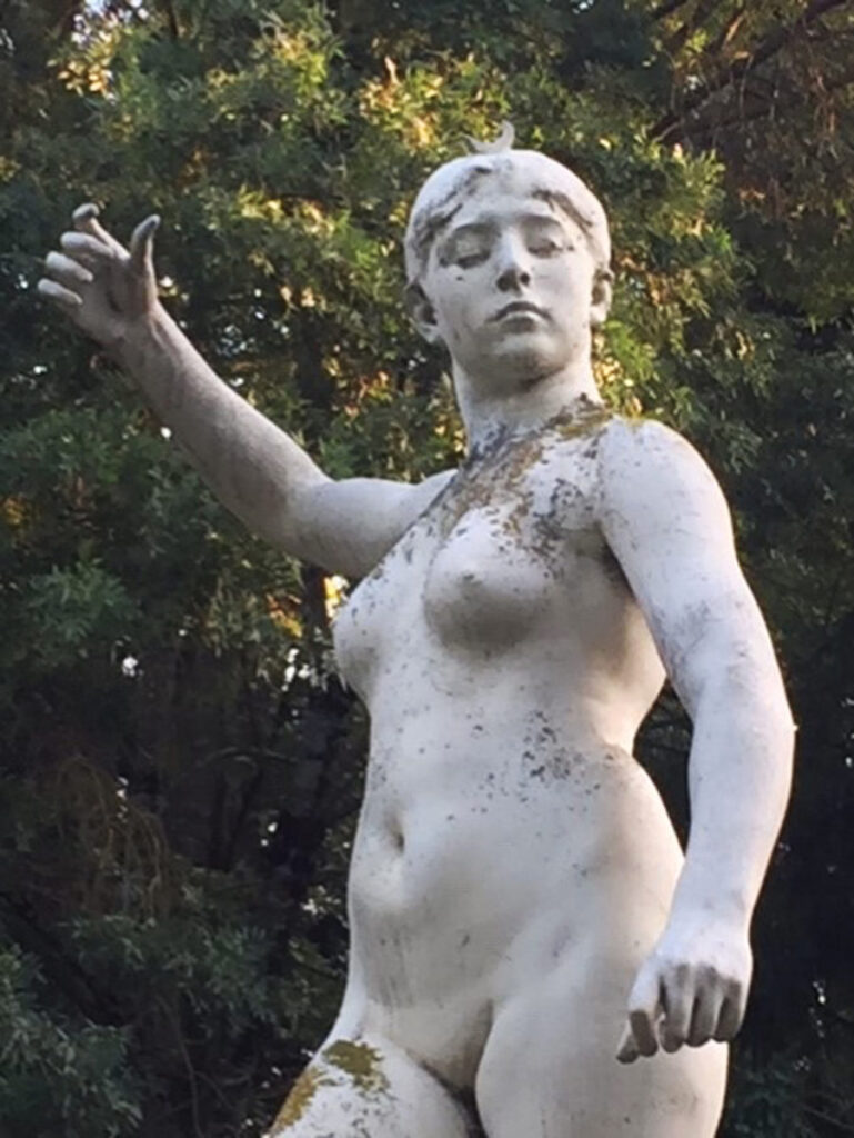 Stone statue of a nude female with raised arm, moss growth, surrounded by dense greenery and trees.