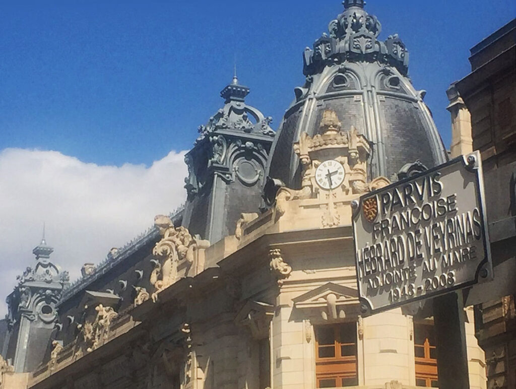 Historic building with ornate stone carvings, clock, and sign reading "Parvis Françoise Hébrard de Veyrinas Adjoint au Maire 1935-2008" against blue sky.