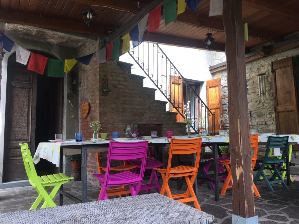 An open-air dining area built of stone, brick, and wood features a long table surrounded by colorful chairs and Buddhist prayer flags overhead at the Il Casale Retreat Center in Merrizo, Italy.