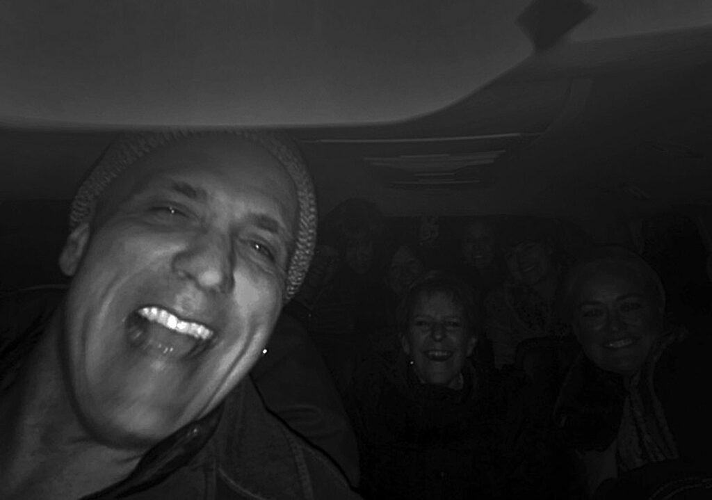 Smiling man with beanie in the foreground, dimly lit vehicle interior, several people in back.