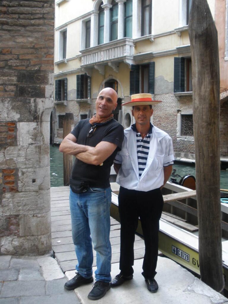 Inner Space host, Ralph Williams, stands next to a Venetian gondolier who's wearing a black & white striped shirt and a classic straw hat with a red band along a canal in Venice, Italy.