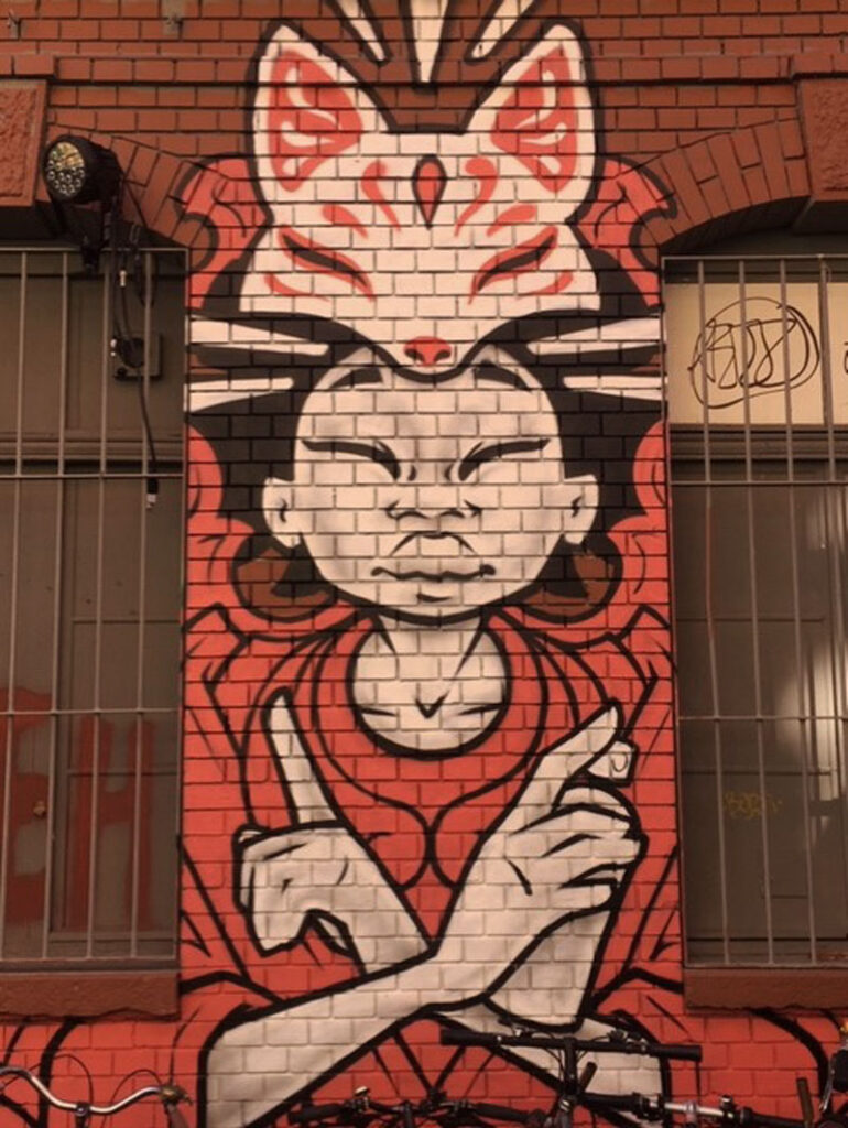 A mural of a person in red with closed eyes, arms crossed, and a fox mask on a brick wall with grates.