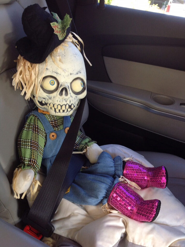 Halloween doll with a skull head and bulging eyes, dressed in plaid shirt, overalls, and purple boots.