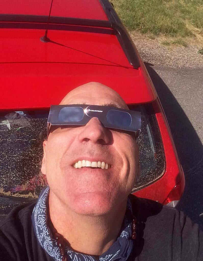 Man in bandana and protective glasses looking up, red car in background, outdoors.