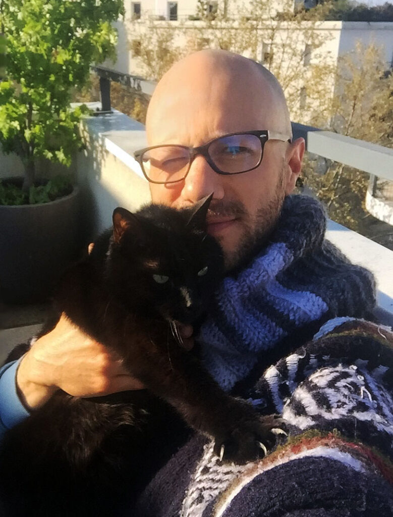 Selfie of Antoine Gee wearing a poncho while holding his black cat, Minou, in Lyon, France.