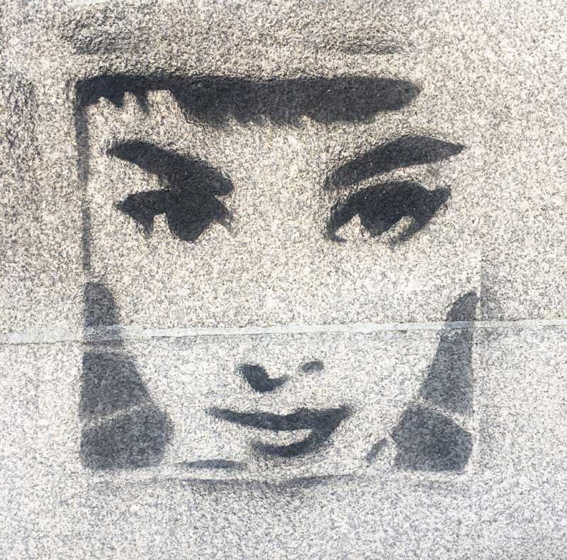 Black and white stencil of a stylized female face with prominent eyes and lips on rough concrete.