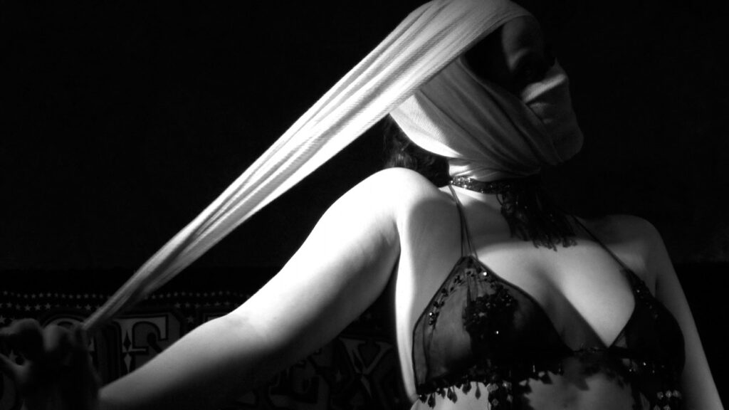 Black & white photo by Grace McEvoy of a female burlesque performer in a see-through top with her head wrapped in white material and one arm pulling on the cloth.