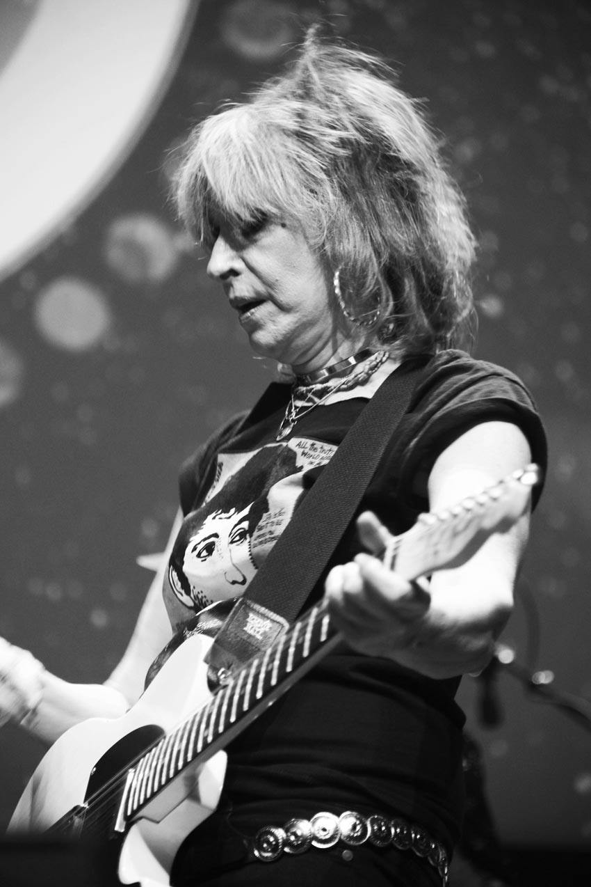 Black & white upper-body photo by Grace McEvoy of musical artist, Chrissie Hynde, playing her guitar on stage.