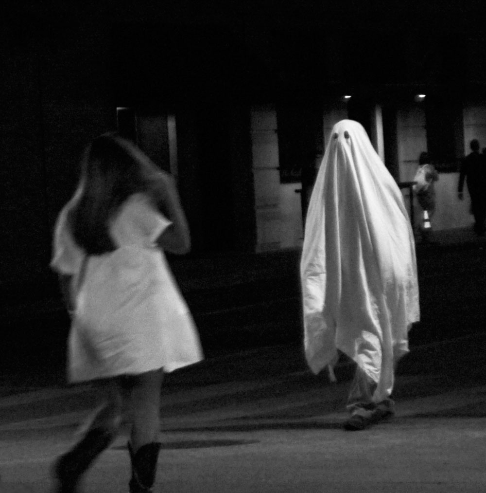 Black & white street photo by Grace McEvoy of a man in a white sheet dressed as a Halloween ghost and a young woman in a short dress and boots walking toward him.