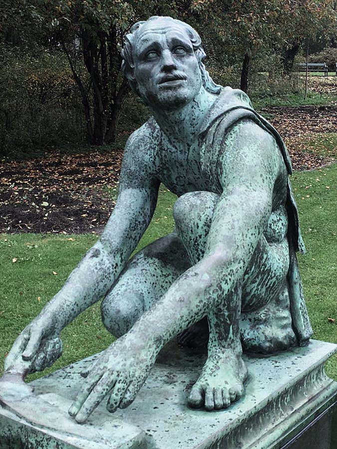 Metal-cast sculpture in Ørsteds Park in Copenhagan of the Greco-Roman mythological slave, Arrotino, crouching and sharpening his knife while looking worried.