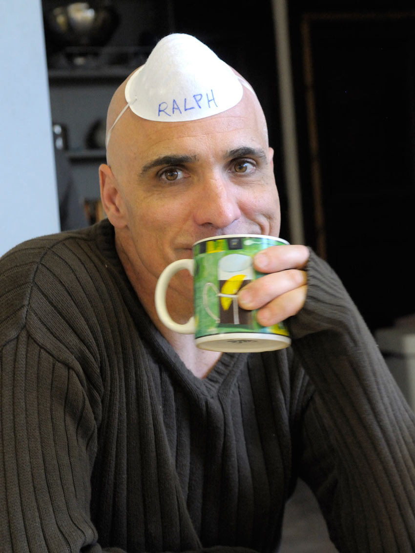 Ralph Williams wears a white filtration mask that says "RALPH" on top of his head while sitting and sipping coffee at the Kagyu Samye Dzong Centre in London, 2009.