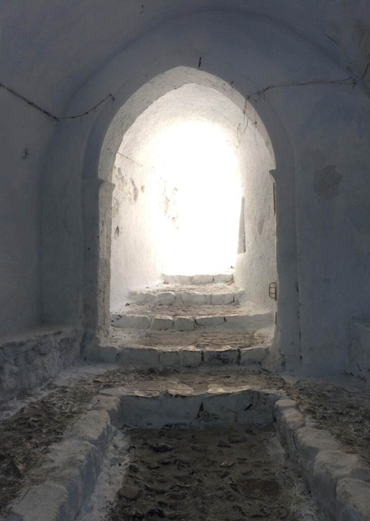 Tucked beneath the Church of the Agia Triada (Holy Trinity) in Pyrgos Village on the Isle of Santorini in Greece is a beautiful monastic chamber with whitewashed walls and white stone steps leading upward into blinding white daylight.