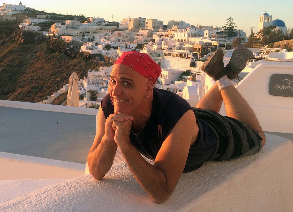 Ralph Williams smiles in a red bandana while lying on his stomach on a white ledge in Santorini, Greece.