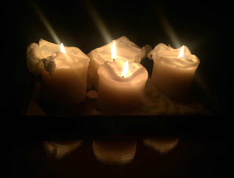 Four white candle-pillars burn on a rectangular plate in a black room.