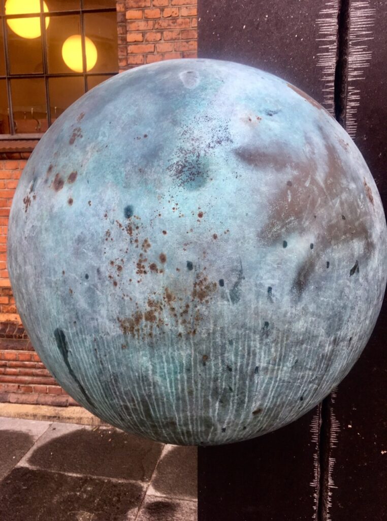 A blue orb sculpture with a stained patina appears to float in space (like Earth) in an outdoor area in Copenhagen.