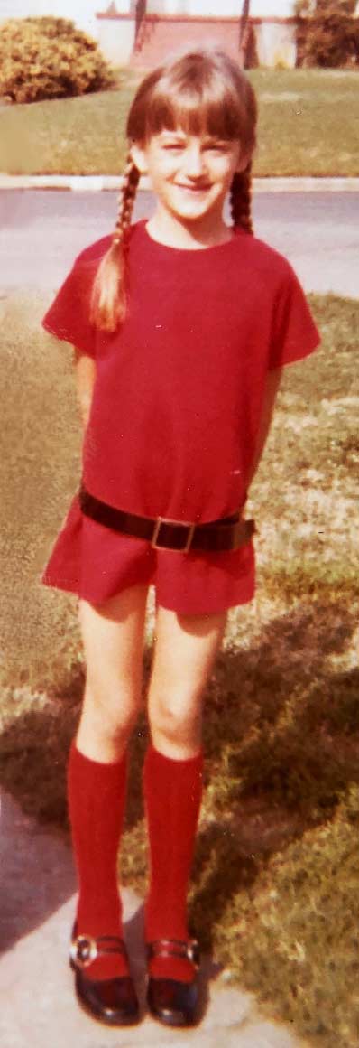 Dana Brock grins at age 10 while wearing a red dress, black belt, and braids.