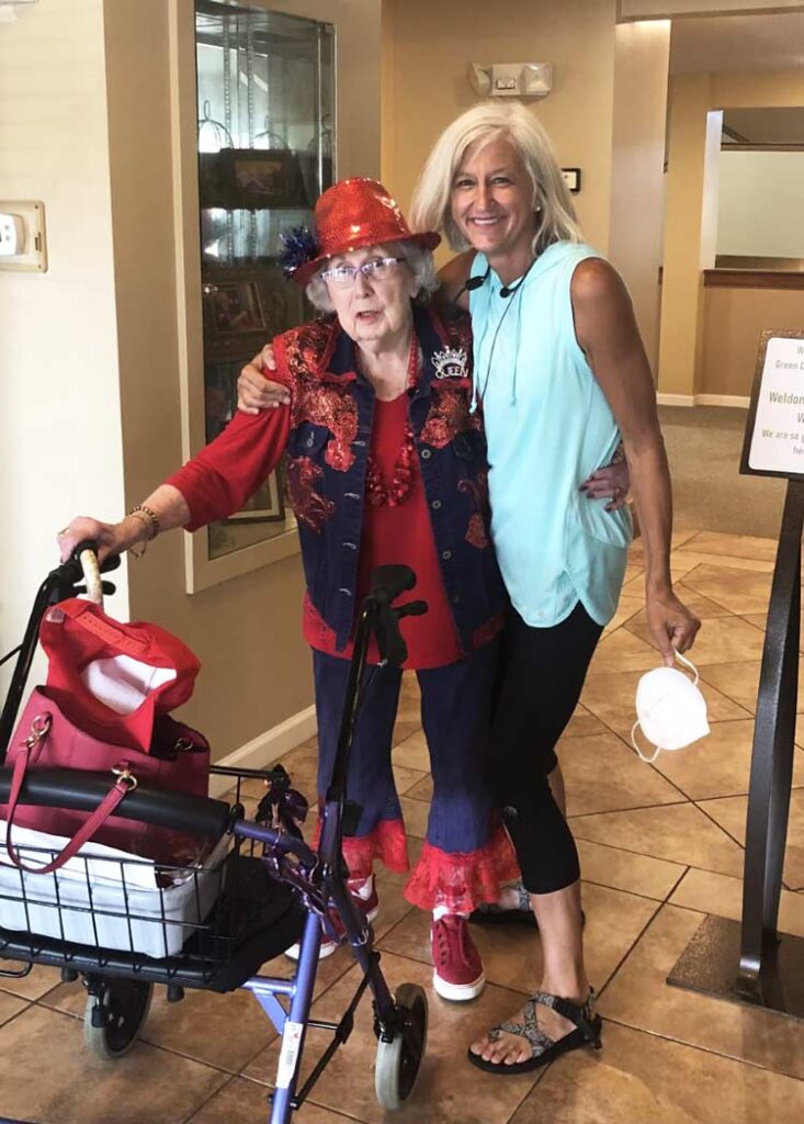 Dana Brock stands arm-in-arm with an elderly woman at an assisted living facility who's wearing a playful red outfit.