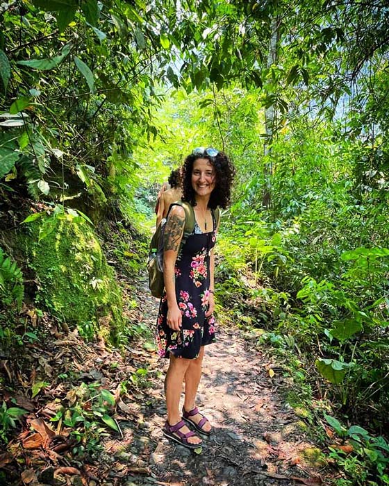 Smiling photo of plant medicine specialist, Misa Miller, in a black floral dress and sandals as she walks a wooded trail.