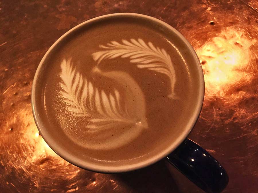 Looking down at a freshly-made cappucino with two leafy shapes in the foam as it sits on a copper countertop.