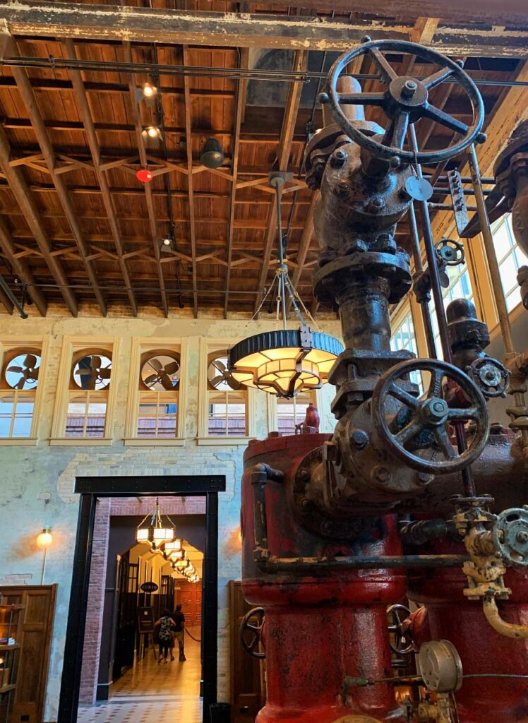 Interior view of the lobby of the Emma Hotel which includes large industrial machinery (for brewing), a raw-wood ceiling, and large ventilation fans in the upper windows.