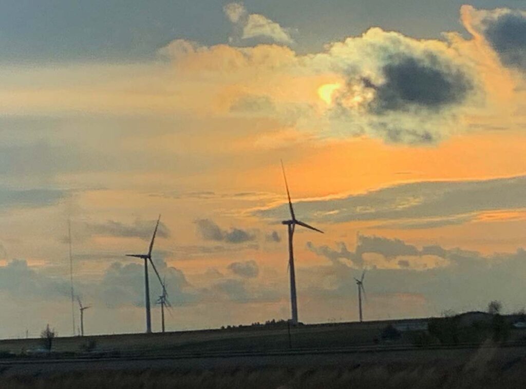 Wind turbines in a flat field under a colorful sunset sky with scattered clouds in the background.