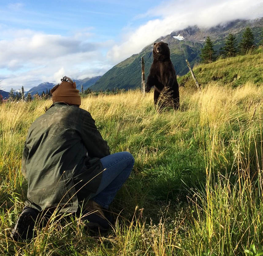 While touring remote Alaska, a female photographer hunkers in the grass to catch a shot of a bear that's standing in front of her.