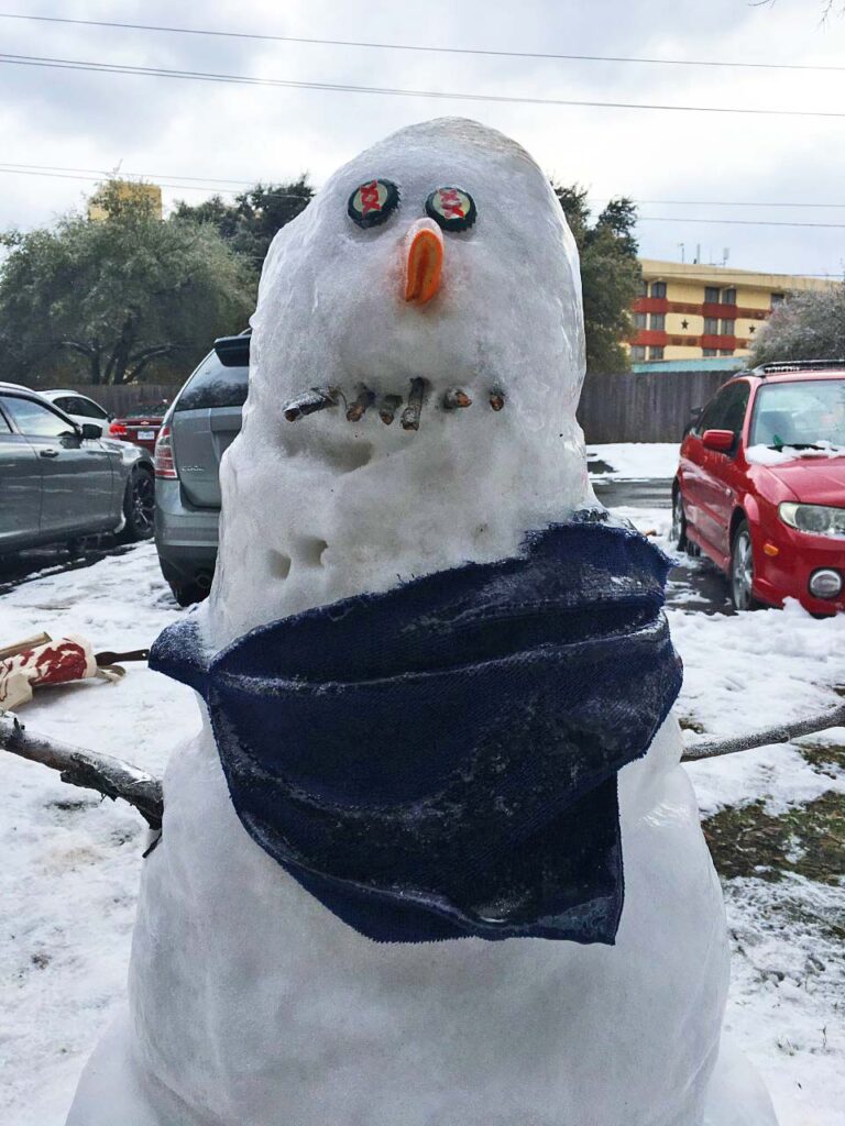 Snowman with bottle cap eyes, carrot nose, twig mouth, scarf, parked cars, snow-covered area.