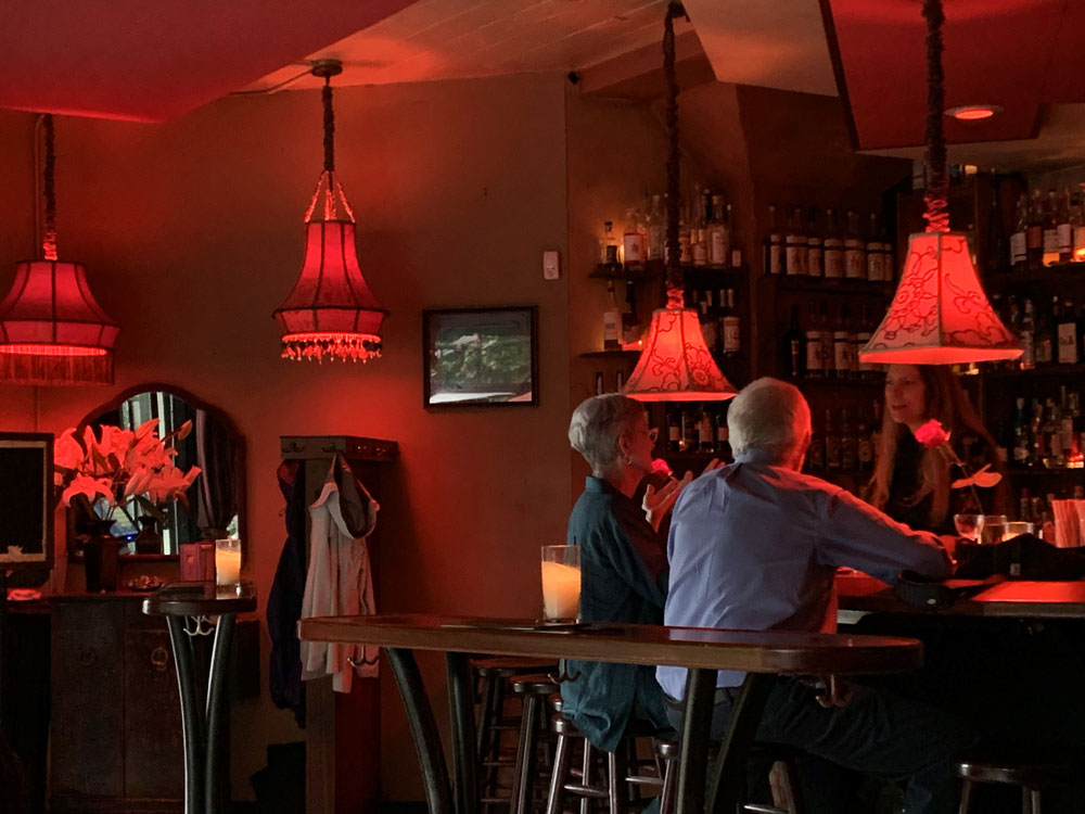 Dimly lit bar with red lanterns, three people talking at counter, shelves with bottles in background