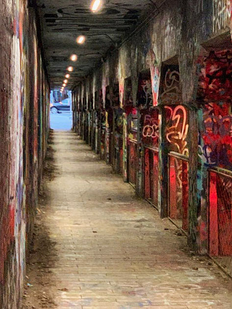 Narrow, covered corridor with graffiti-covered columns and brick-paved floor, illuminated overhead.