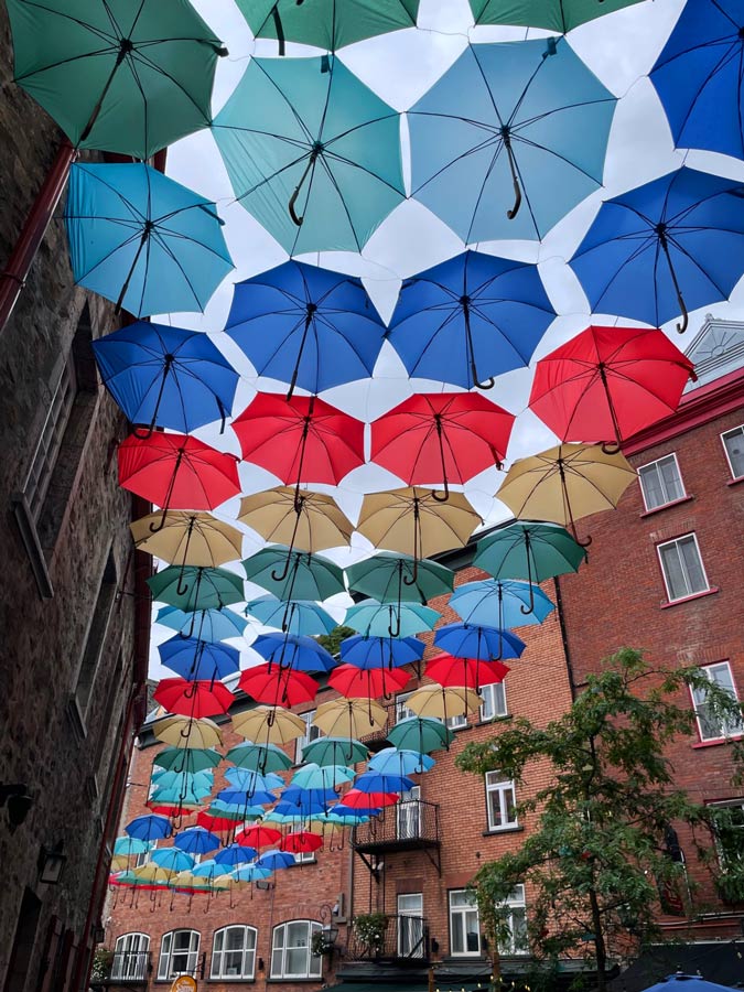 Colorful umbrellas suspended over a narrow alley with brick and stone buildings and greenery.