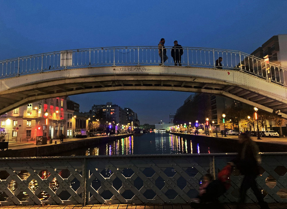 Nighttime city scene with a lit-up river, white pedestrian bridge, and people walking along the waterfront.