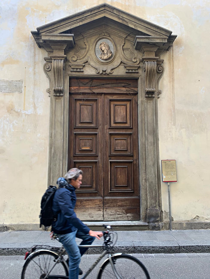 Person cycling past large carved wooden door with ornate stone archway and plaque on wall.