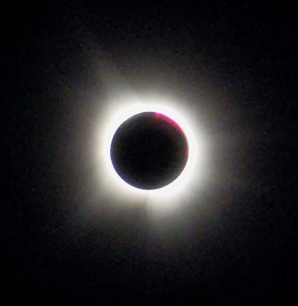 Solar eclipse with the moon covering the sun and the bright white light of the sun emanating outward, set against a pitch black sky.